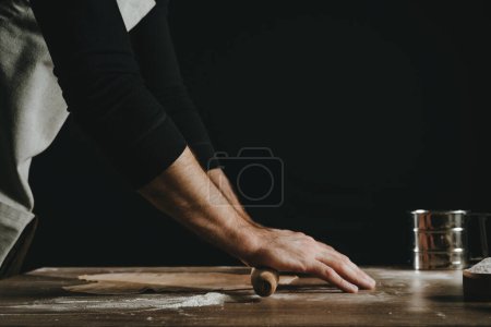Téléchargez les photos : Homme méconnaissable déroulant la pâte avec un rouleau à pâtisserie sur fond noir. Gros plan des mains masculines préparant les nouilles maison. Concentration sélective. - en image libre de droit