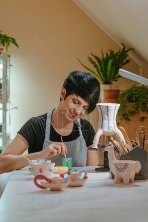 Foto de Joven artesana pintando un jarrón hecho a mano sentado en su taller. Enfoque selectivo. - Imagen libre de derechos