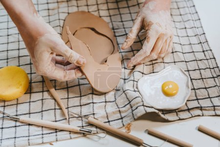 Photo for Young woman potter carving a fried eggs shape plate from clay in her workshop. Close-up of female hands. Selective focus. - Royalty Free Image
