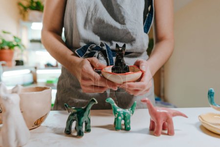Photo for Young woman ceramist holding small colorful ceramic dinosaur figurines in the workshop. Close up, selective focus. - Royalty Free Image