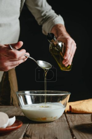 Téléchargez les photos : Homme méconnaissable dans un tablier ajoutant de l'huile végétale à un bol en verre avec du lait pendant la cuisson. Concentration sélective. - en image libre de droit