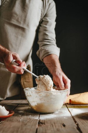 Photo for Unrecognizable man in an apron kneading the dough with a spoon on a wooden table. Black background. Selective focus. - Royalty Free Image