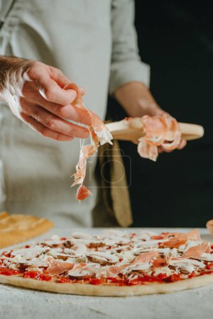 Photo for Close-up of chef preparing pizza putting ham prosciutto slices on the dough. Selective focus. - Royalty Free Image