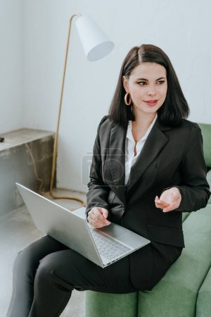 Téléchargez les photos : Jeune femme d'affaires souriante dans un costume avec un ordinateur portable sur ses genoux assis sur un canapé vert parler. En arrière-plan se trouve un mur blanc et un lampadaire blanc. - en image libre de droit