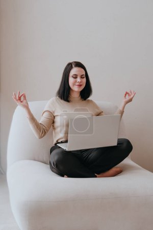 Téléchargez les photos : Jeune femme brune souriante aux yeux fermés dans des vêtements d'affaires assis pieds nus en position lotus avec un ordinateur portable à l'intérieur. Équilibre entre travail et santé mentale. - en image libre de droit