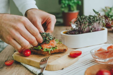 Photo for Closeup of males hands making sandwich with salmon fish, radish sprouts microgreen and tomato on wooden background. Selective focus. - Royalty Free Image