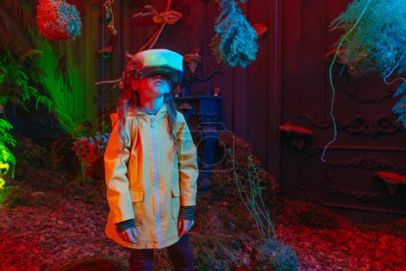 Photo for Little girl wearing luminous virtual reality headset standing in the room with moss, fern, branches in neon lights. Augmented reality game in natural conditions. Concept of connection of the future technology and nature. Selective focus. - Royalty Free Image