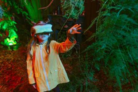 Photo for Little girl wearing luminous virtual reality headset standing in the room with moss, fern, branches in red neon lights. Concept of connection of the future technology and nature. Selective focus. - Royalty Free Image