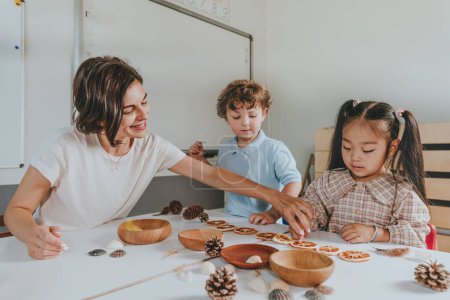 Photo for Boy and girl playing with pine cones, dried citrus and seashells together with teacher in a kindergarten. Early education with natural materials - Royalty Free Image