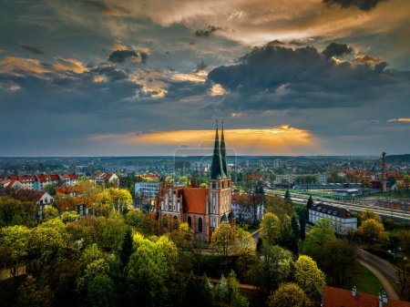 Foto de Olsztyn Iglesia de la Guarnición de Nuestra Señora Reina de Polonia, parque fuera del castillo desde la vista de un pájaro. Por encima de la iglesia, los rayos del sol poniente rompiendo las nubes. Época del año - primavera. - Imagen libre de derechos