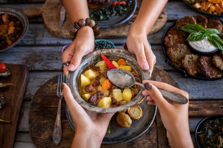Foto de Meat stew with vegetables at the hands of two women. Orman kebabi . Cooked foods in the copper plate or pot. - Imagen libre de derechos