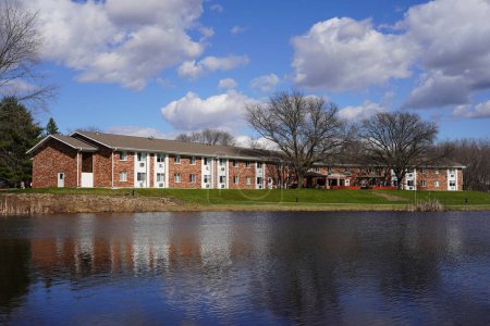 Téléchargez les photos : Mauston, Wisconsin États-Unis - 13 avril 2024 : vue du paysage des immeubles d'appartements en brique sur le bord d'un lac par une journée nuageuse. - en image libre de droit