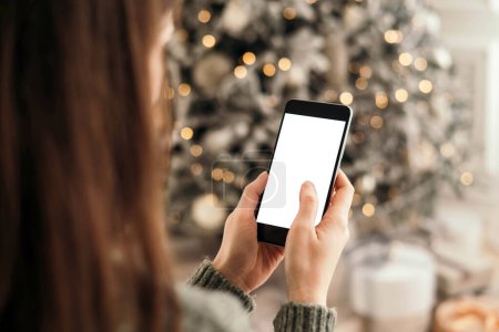 Photo for Christmas mockup with space for text. christmas advertising, app template. hands holding phone with empty screen on background of beautiful christmas tree with lights. - Royalty Free Image