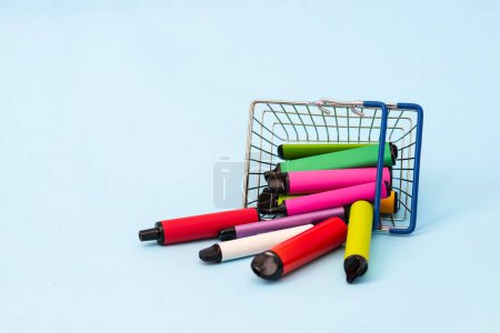 Téléchargez les photos : Un ensemble de bâtons multicolores dispositifs de vapotage jetables, cigarettes électroniques dans un panier sur un fond bleu - en image libre de droit