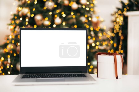 Téléchargez les photos : Vue de face de l'ordinateur portable avec écran blanc dans la maison avec arbre de Noël lumière chaude et boîte cadeau. Rabais Black Friday. Achats en ligne. Cadeaux pour la nouvelle année et Noël - en image libre de droit