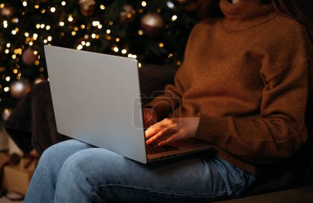 Téléchargez les photos : Achats en ligne de Noël la nuit, promotions des ventes et des rabais pendant les vacances d'hiver, achats en ligne à la maison. Mains féminines sur l'ordinateur portable et les guirlandes. - en image libre de droit