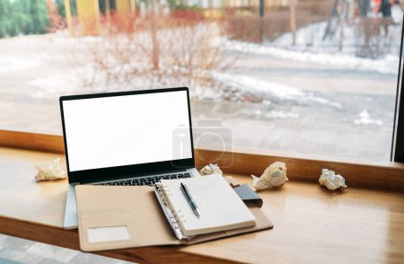 Computer-Laptop mit weißem Blanko-Bildschirm, zusammengestellt mit Notizbuch und auf dem modernen Holztisch mit Fenstern im Café als Hintergrund