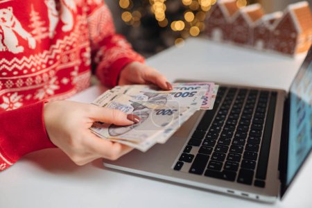Téléchargez les photos : Femme comptant les factures de hryvnia ukrainienne, à l'aide d'un ordinateur portable. Fille compter les cadeaux de Noël. - en image libre de droit
