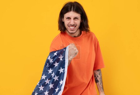 Porträt eines motivierten Mannes aus dem Nahen Osten, der die US-Flagge in die Kamera hält. Feiertag, eingefleischter Fan mit langen Haaren und Tätowierung auf gelbem Hintergrund