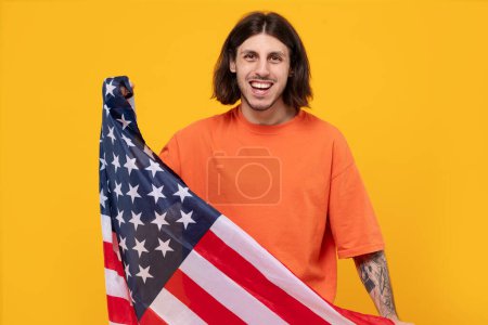 Porträt eines motivierten Mannes aus dem Nahen Osten, der die US-Flagge in die Kamera hält. Feiertag, eingefleischter Fan mit langen Haaren und Tätowierung auf gelbem Hintergrund