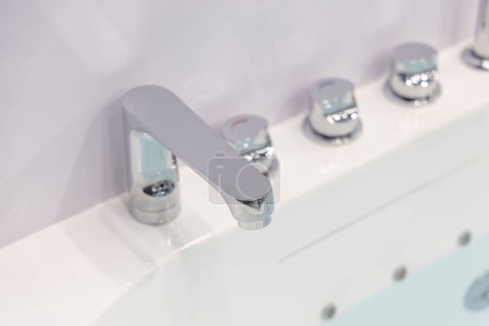Modern chrome faucet and water taps on a white acrylic bathtub. All parts are made of chromed metal, reflecting the quality and style of the sanitary equipment.