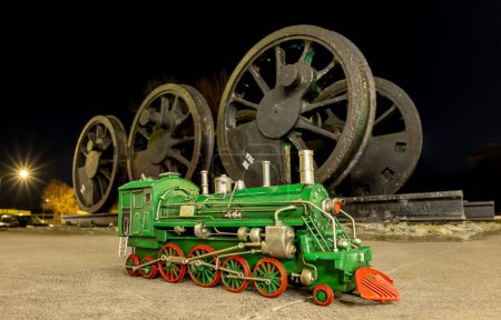 Foto de Modelo de locomotora de vapor verde con ruedas rojas delante de un conjunto de ruedas de locomotora de vapor antiguas. - Imagen libre de derechos