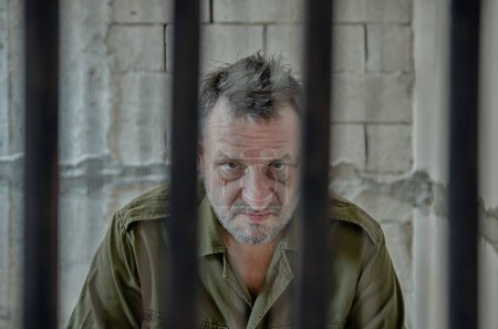 Foto de Un anciano con una camisa verde del ejército tras las rejas de la prisión. - Imagen libre de derechos