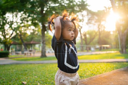 Foto de Adorable poco asiático chica caminando en verde hierba público parque mañana amanecer - Imagen libre de derechos