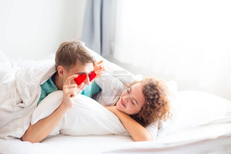 Téléchargez les photos : Couple d'amour couché sur le lit sous la couverture tenant les coeurs rouges, vie romantique heureuse - en image libre de droit