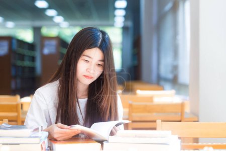 Téléchargez les photos : Heureux asiatique étudiant jeune femme penser avec livre dans bibliothèque assis sur bois table - en image libre de droit
