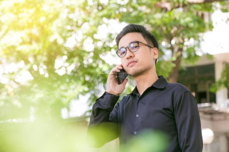Photo for Happy young handsome man stand under tree outdoors and using smartphone for business talk, Glasses man - Royalty Free Image