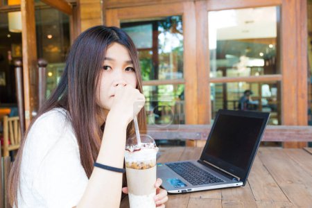 Téléchargez les photos : Heureuse femme souriante travaillant avec un ordinateur portable et buvant du café latte froid sur une table en bois au café - en image libre de droit