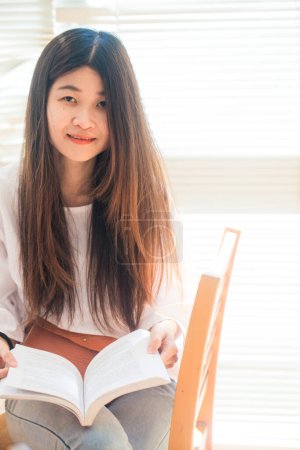 Téléchargez les photos : Heureux asiatique étudiant jeune femme penser avec livre dans bibliothèque assis sur bois table - en image libre de droit
