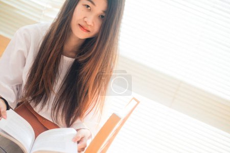 Foto de Feliz asiático estudiante joven mujer pensando con libro en biblioteca sentado en madera mesa - Imagen libre de derechos