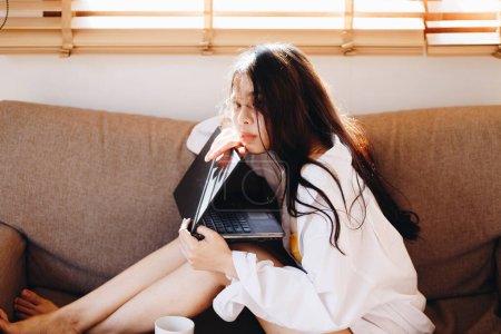 Foto de Negocios mujer asiática acostada en sofá uso ordenador portátil en casa acogedora mañana despertar el trabajo desde cualquier lugar - Imagen libre de derechos