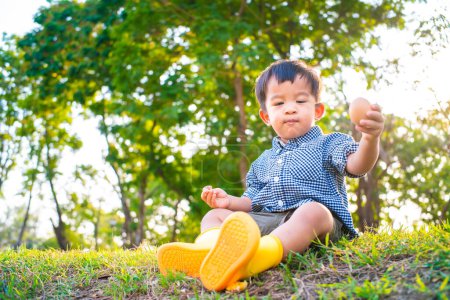 Foto de Feliz asiático chico con huevo de Pascua en verde hierba césped amarillo bota zapato en ciudad parque puesta de sol luz - Imagen libre de derechos