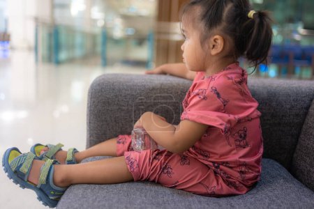 Téléchargez les photos : Adorable fille asiatique de 2 ans assis sur un canapé confortable à l'hôpital brouillé concept de soins de santé de fond - en image libre de droit