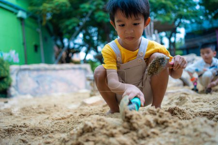 Foto de Jardín de infancia asiático 5 años de edad jugando en sandbox divertirse en el patio de recreo en arenero. Actividades creativas al aire libre - Imagen libre de derechos