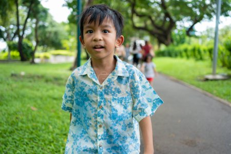 Téléchargez les photos : Heureux asiatique préscolaire garçon marche dans vert tropical arbre forêt dans ville parc loisirs de plein air - en image libre de droit