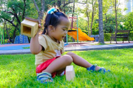 Photo for Adorable little toddler girl play wooden house toy sitting on green grass in city public park morning sun light - Royalty Free Image