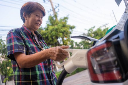 Téléchargez les photos : Asiatique mature femme tenir câble de charge à la voiture électrique à la station, respectueux de l'environnement zéro émission - en image libre de droit