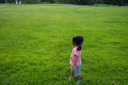 Photo for Pretty toddler asian girl enjoying walk and run on green grass in city park sunset light - Royalty Free Image