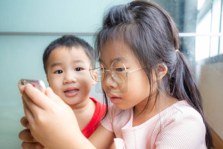 Foto de Hermana y hermano niño jugando smartphone en la habitación hermano mira hermana - Imagen libre de derechos