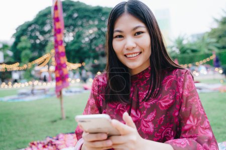 Téléchargez les photos : Belles femmes d'affaires asiatiques utilisent un message texte smartphone assis sur l'herbe dans le parc de fête arrière-plan extérieur - en image libre de droit