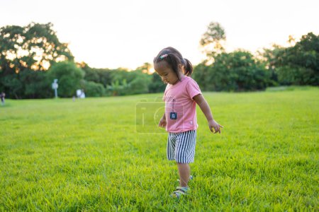 Téléchargez les photos : Adorable tout-petit fille asiatique profiter de la promenade et courir sur l'herbe verte dans le parc de la ville coucher de soleil fille lumière avec la nature - en image libre de droit