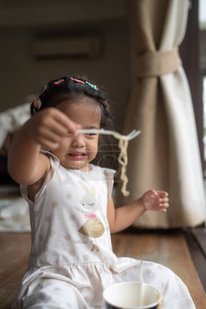 Foto de Adorable poco asiático chica disfrutando comer fideos en acogedor casa - Imagen libre de derechos