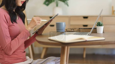 Téléchargez les photos : Jeune femme d'affaires écrit des notes de données de projet d'entreprise dans la tablette et le travail sur ordinateur portable à la maison. - en image libre de droit