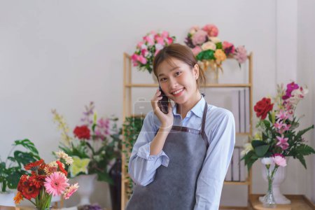 Téléchargez les photos : Concept de fleurisme, Fleuriste femme parlant avec le client sur smartphone tout en se tenant debout dans le magasin de fleurs. - en image libre de droit