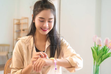 Téléchargez les photos : Jeunes femmes asiatiques portant un bracelet avec bonheur après avoir enfilé des perles colorées sur le fil pour faire des accessoires faits à la main tout en faisant un mode de vie de passe-temps et la création de bijoux artisanat à la maison atelier. - en image libre de droit