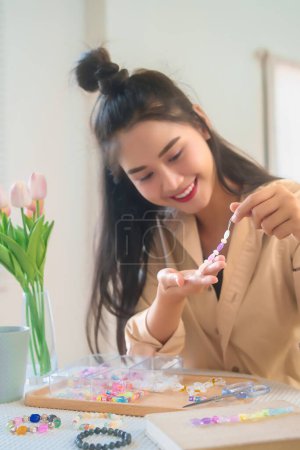 Téléchargez les photos : Jeunes femmes asiatiques ficelant des perles sur le fil et choisissant des perles colorées de la boîte à la fabrication de bracelet à la main tout en passant du temps à créer des bijoux artisanaux et à faire un style de vie de passe-temps à la maison. - en image libre de droit